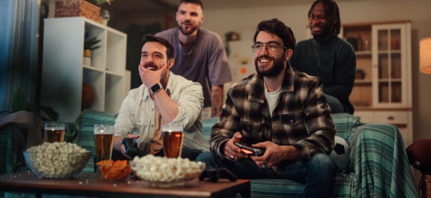 Four men sitting on a couch holding game controllers, with a bowl of snacks in front of them